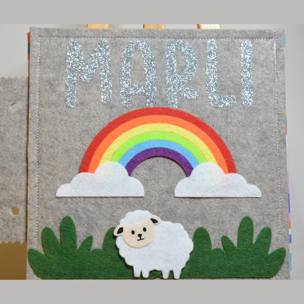 Felt busy book cover: a rainbow over layered clouds with a sheep in the grass, and a name cut in silver glitter