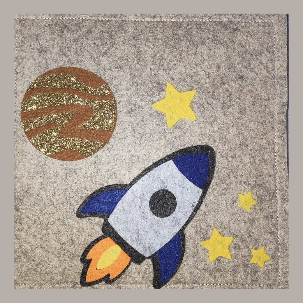 Felt busy book cover: a rocket climbing past a gold-banded planet and yellow stars, on grey felt